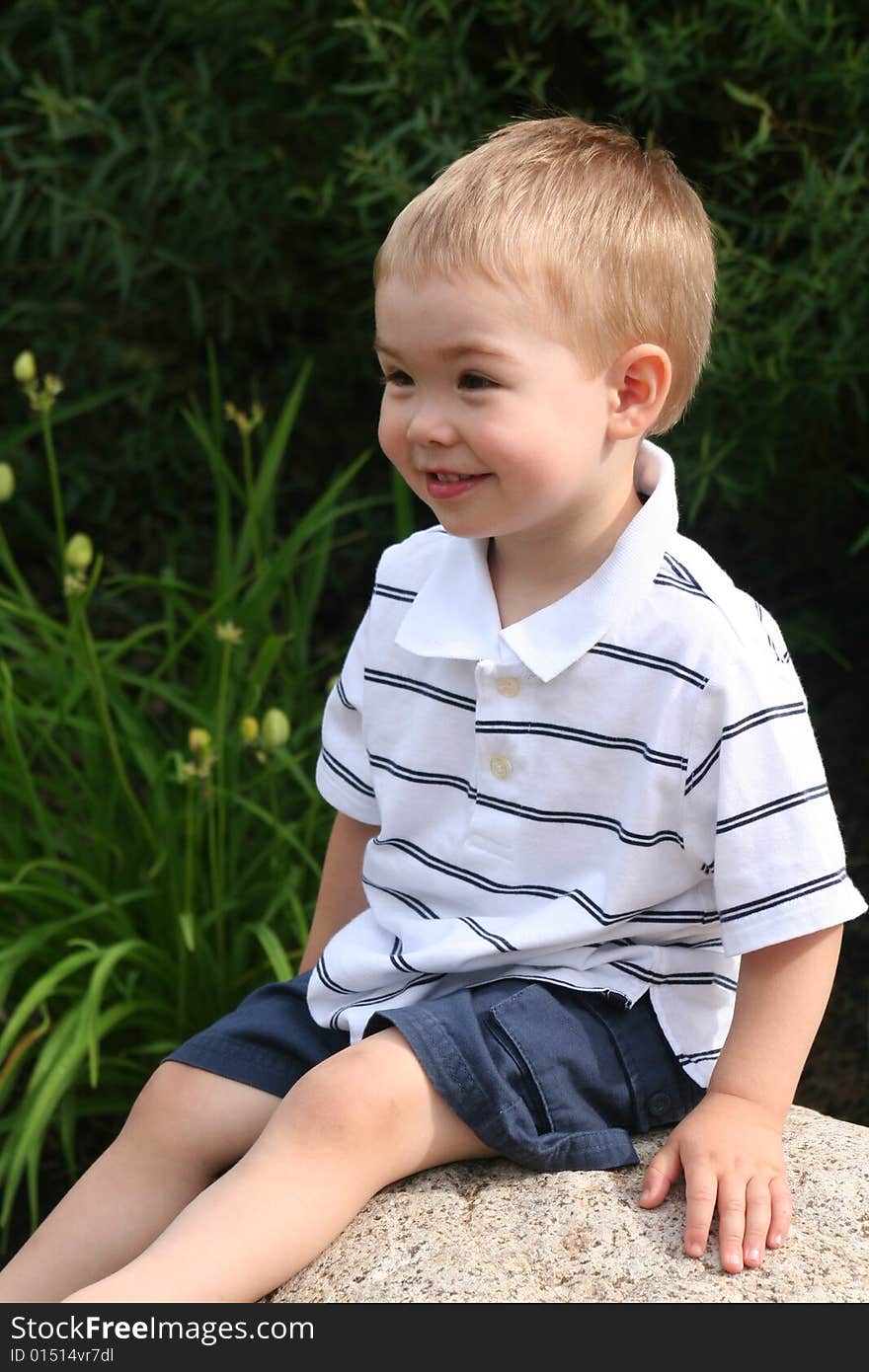 A Toddler boy wearing a striped white golf shirt sitting on a rock looking away from the camera. A Toddler boy wearing a striped white golf shirt sitting on a rock looking away from the camera.