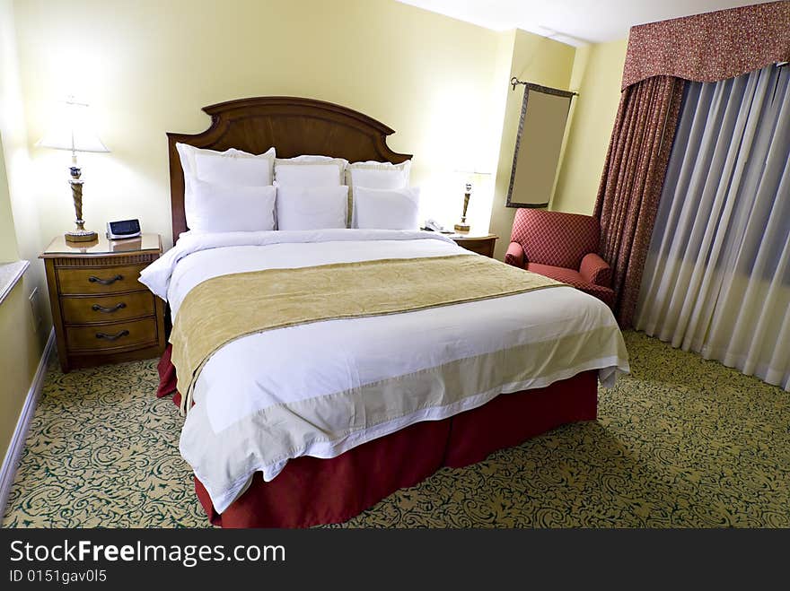 View of large king sized bed with nightstand and lamp and curtained window in background. View of large king sized bed with nightstand and lamp and curtained window in background