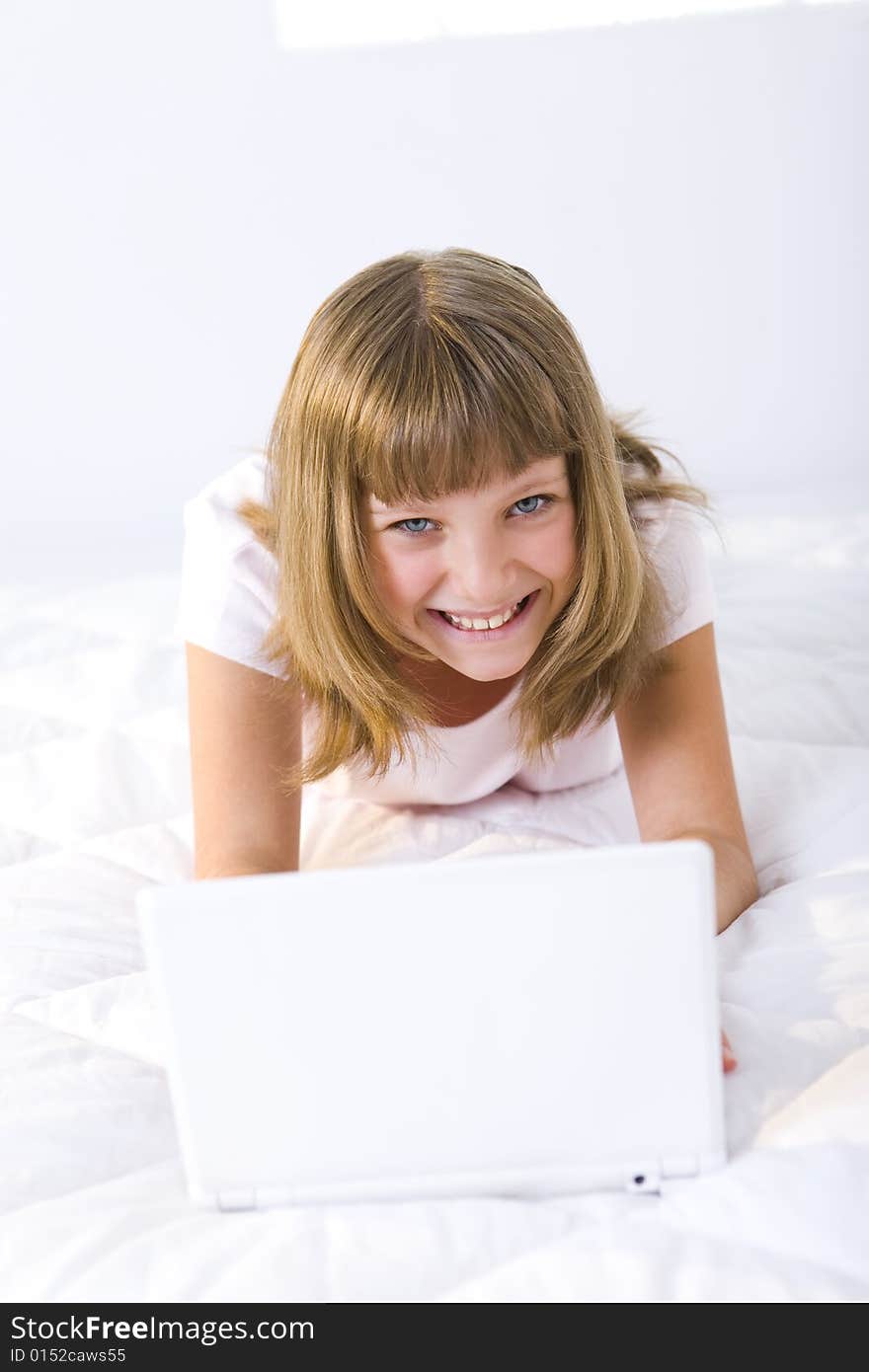 Young smiling girl lying on a bed and does something on laptop. She's looking at camera. Young smiling girl lying on a bed and does something on laptop. She's looking at camera.