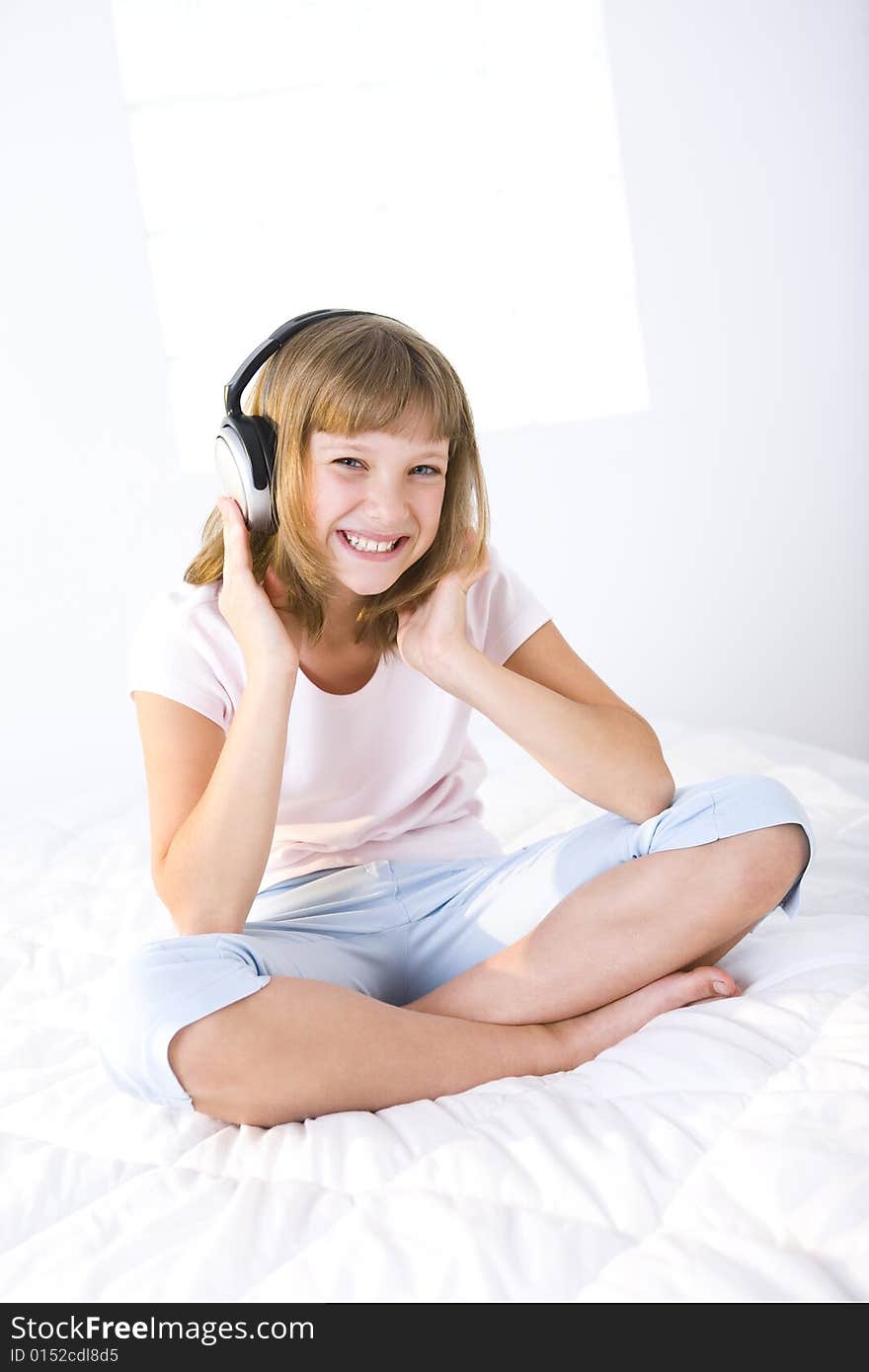 Young smiling girl sitting cross-legged on a bed and listening music by earphones. She's looking at camera. Young smiling girl sitting cross-legged on a bed and listening music by earphones. She's looking at camera.