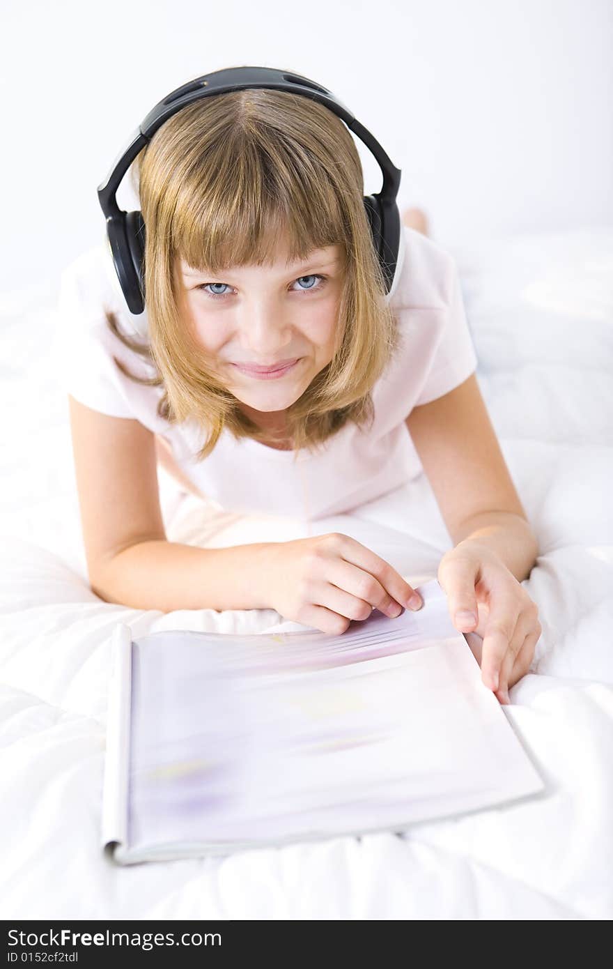 Young smiling girl lying on a bed with earphones. She's browsing a journal. Young smiling girl lying on a bed with earphones. She's browsing a journal.