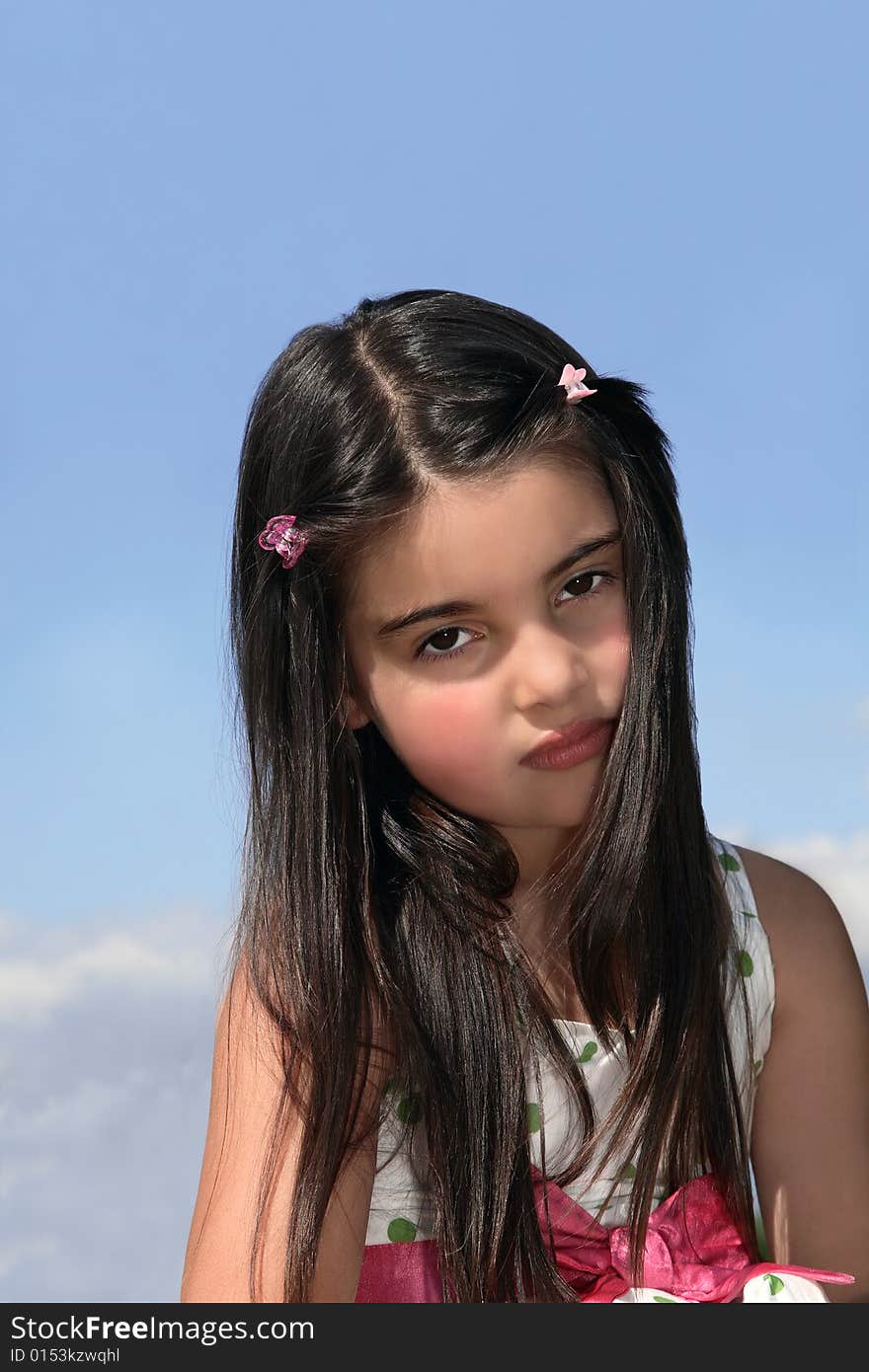 Face of a young little girl with dark hair looking serious. Set against a blue sky with clouds. Face of a young little girl with dark hair looking serious. Set against a blue sky with clouds.