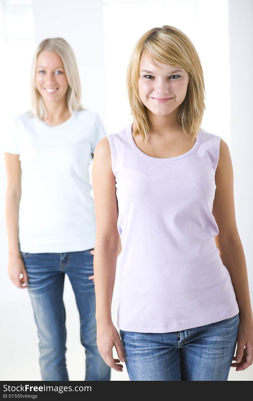 Two smiling girl standing near window and looking at camera. Focused on the girl who standing in front. Front view. Two smiling girl standing near window and looking at camera. Focused on the girl who standing in front. Front view.