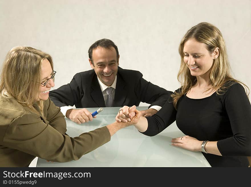 Two women and a man at a table smiling, and the women are arm wrestling. Horizontally framed photo. Two women and a man at a table smiling, and the women are arm wrestling. Horizontally framed photo.