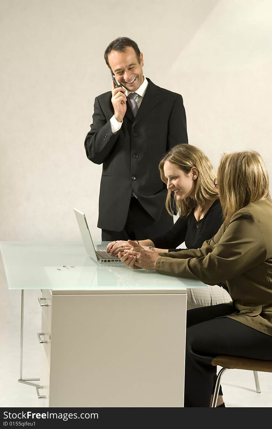 Three people are together in a room at a table. They are all looking at each other and the man is standing up and talking on a cell phone. It appears to be a business meeting and there is a laptop on the table in front of the youngest member. Vertically framed shot. Three people are together in a room at a table. They are all looking at each other and the man is standing up and talking on a cell phone. It appears to be a business meeting and there is a laptop on the table in front of the youngest member. Vertically framed shot.