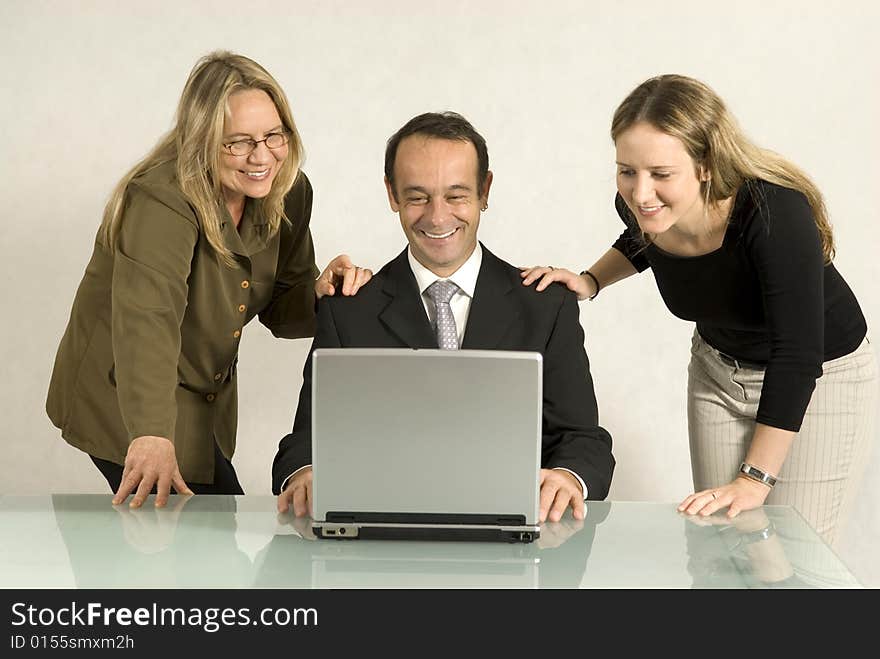 Two women standing over a table where a man is seated looking at a laptop computer. They are all smiling. Two women standing over a table where a man is seated looking at a laptop computer. They are all smiling.