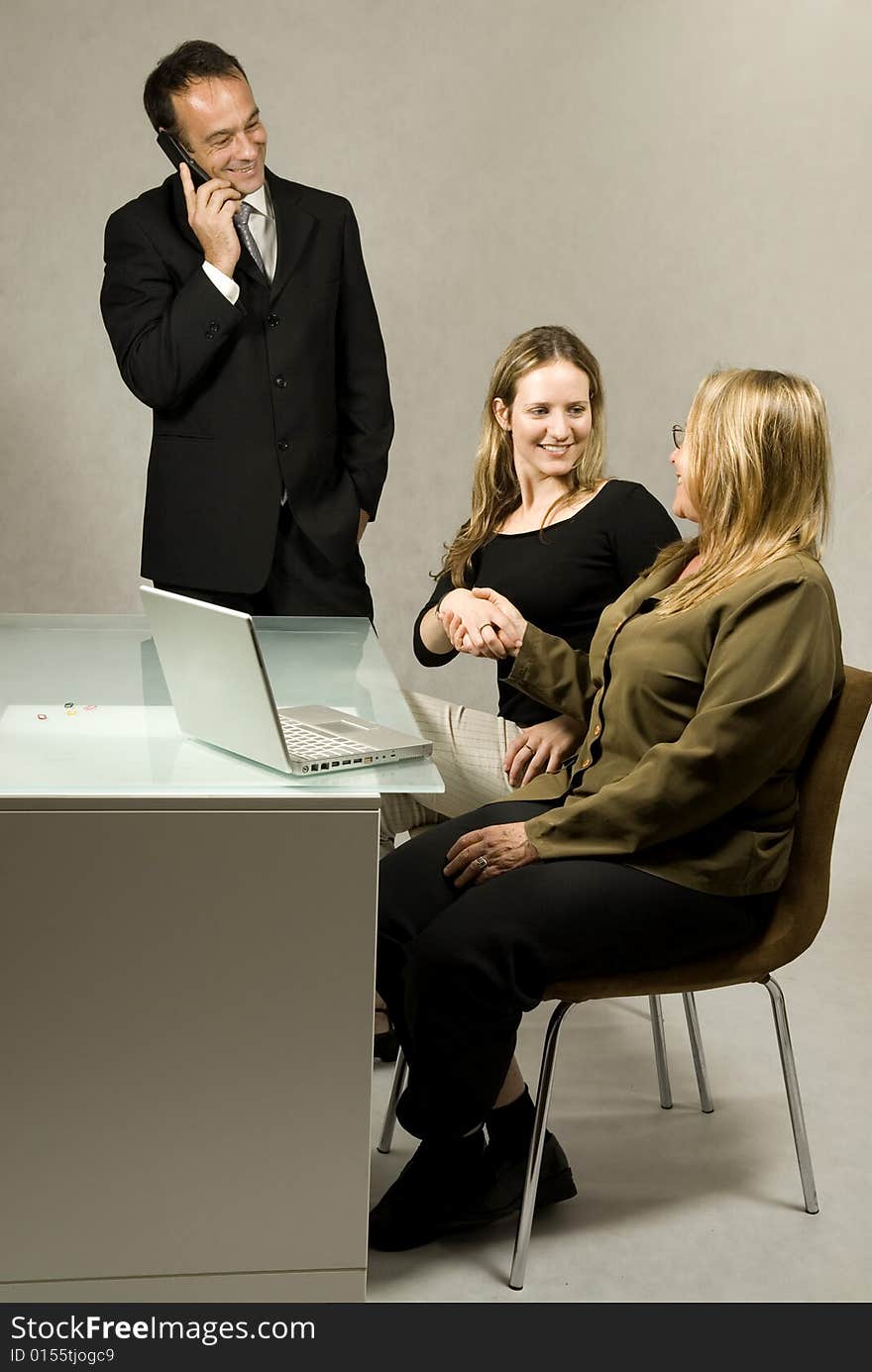 Two women at a desk in front of a laptop shaking hands and smiling, while a man stands on his cell phone smiling. Vertically framed photo. Two women at a desk in front of a laptop shaking hands and smiling, while a man stands on his cell phone smiling. Vertically framed photo.