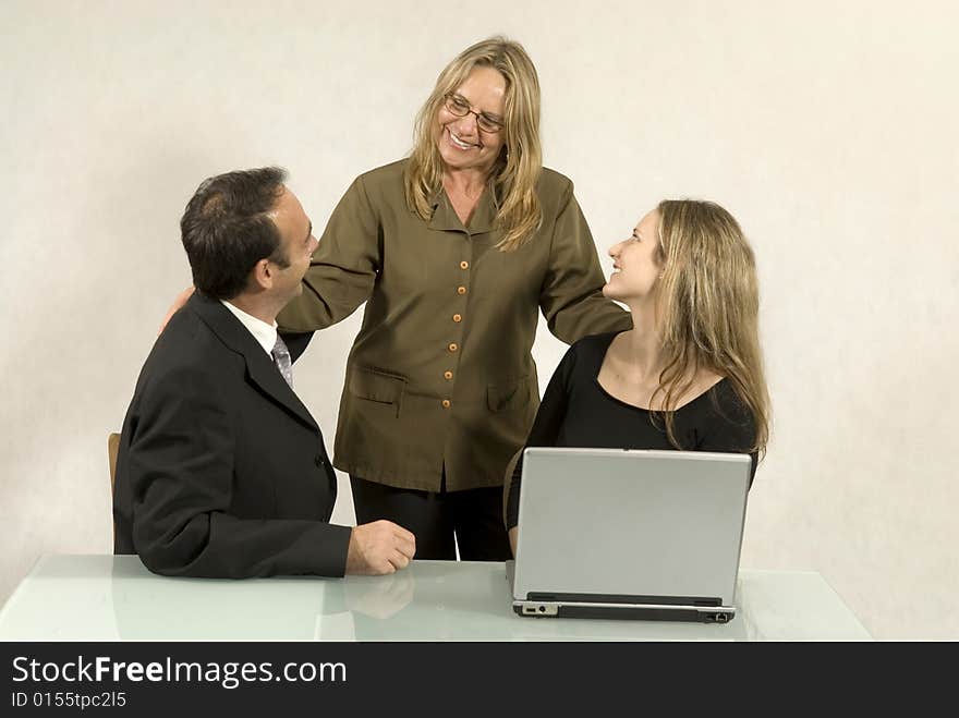 Three people are together in a room at a table. The older woman is standing and the other two are sitting down. They are smiling and looking at each other and there is a laptop on the table in front of the younger woman. Horizontally framed shot. Three people are together in a room at a table. The older woman is standing and the other two are sitting down. They are smiling and looking at each other and there is a laptop on the table in front of the younger woman. Horizontally framed shot.