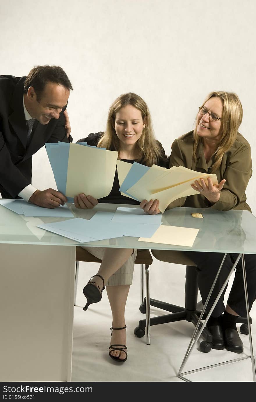 Three people are together in a room at a table. It appears to be a business meeting. They are all looking down at pieces of paper in their hands and smiling. Vertically framed shot. Three people are together in a room at a table. It appears to be a business meeting. They are all looking down at pieces of paper in their hands and smiling. Vertically framed shot.