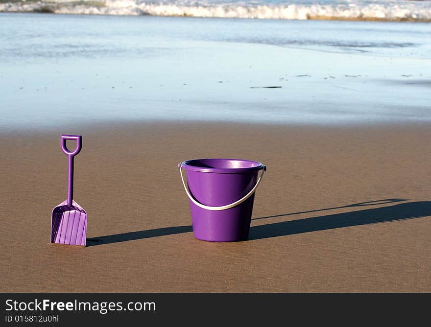 A purple plastic bucket and spade on the beach with the ocean behind them coming in. A purple plastic bucket and spade on the beach with the ocean behind them coming in