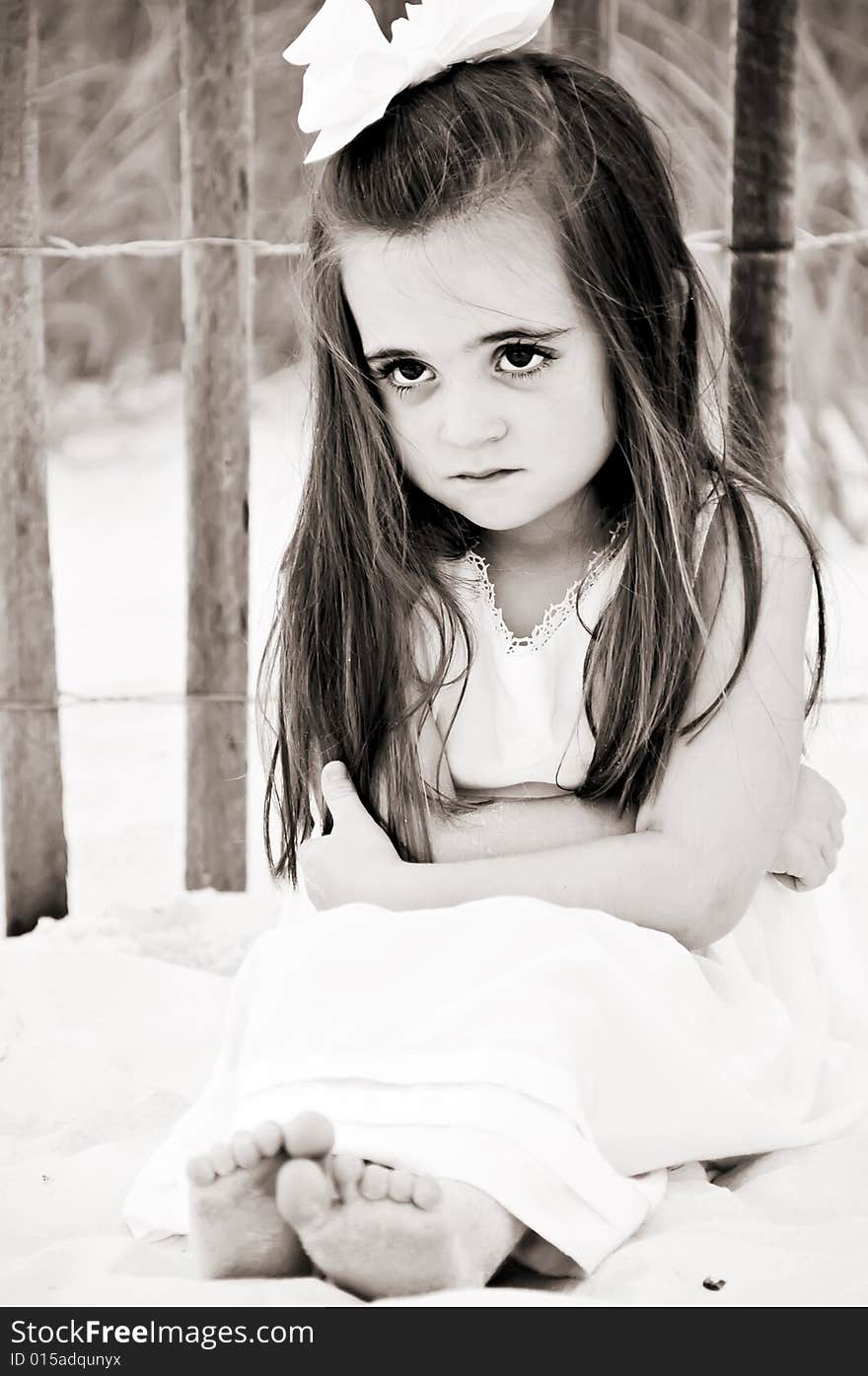 Little girl frowning sitting on the beach by a wooden fence dressed in a white dress with a big bow in her brunette hairin brown tones. Little girl frowning sitting on the beach by a wooden fence dressed in a white dress with a big bow in her brunette hairin brown tones