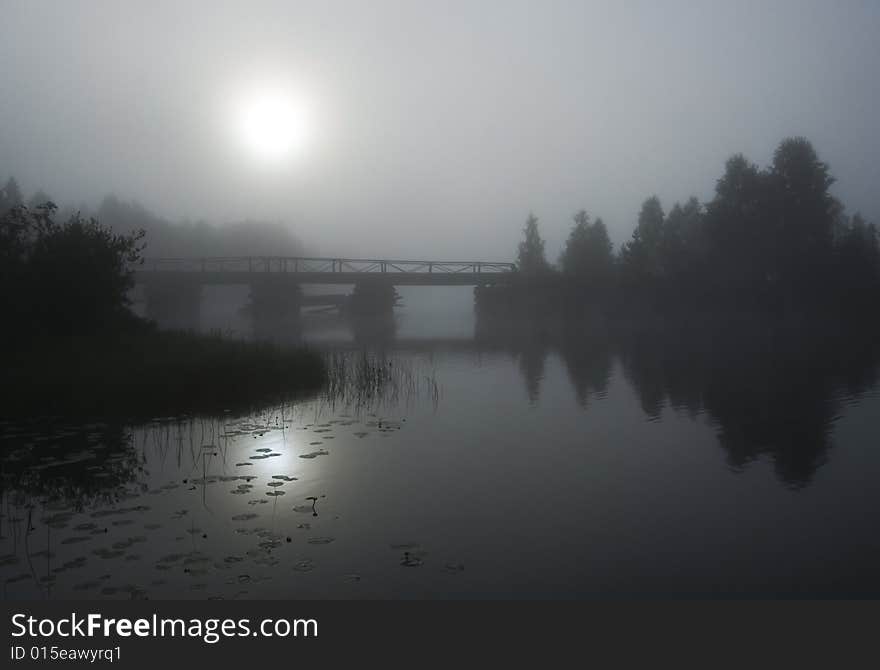 An old wooden bridge looks darkly under the pale sun in the foggy morning