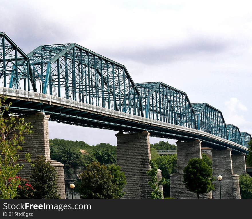 Blue steel bridge with stone columns, trees in background, park below, blue sky. Blue steel bridge with stone columns, trees in background, park below, blue sky