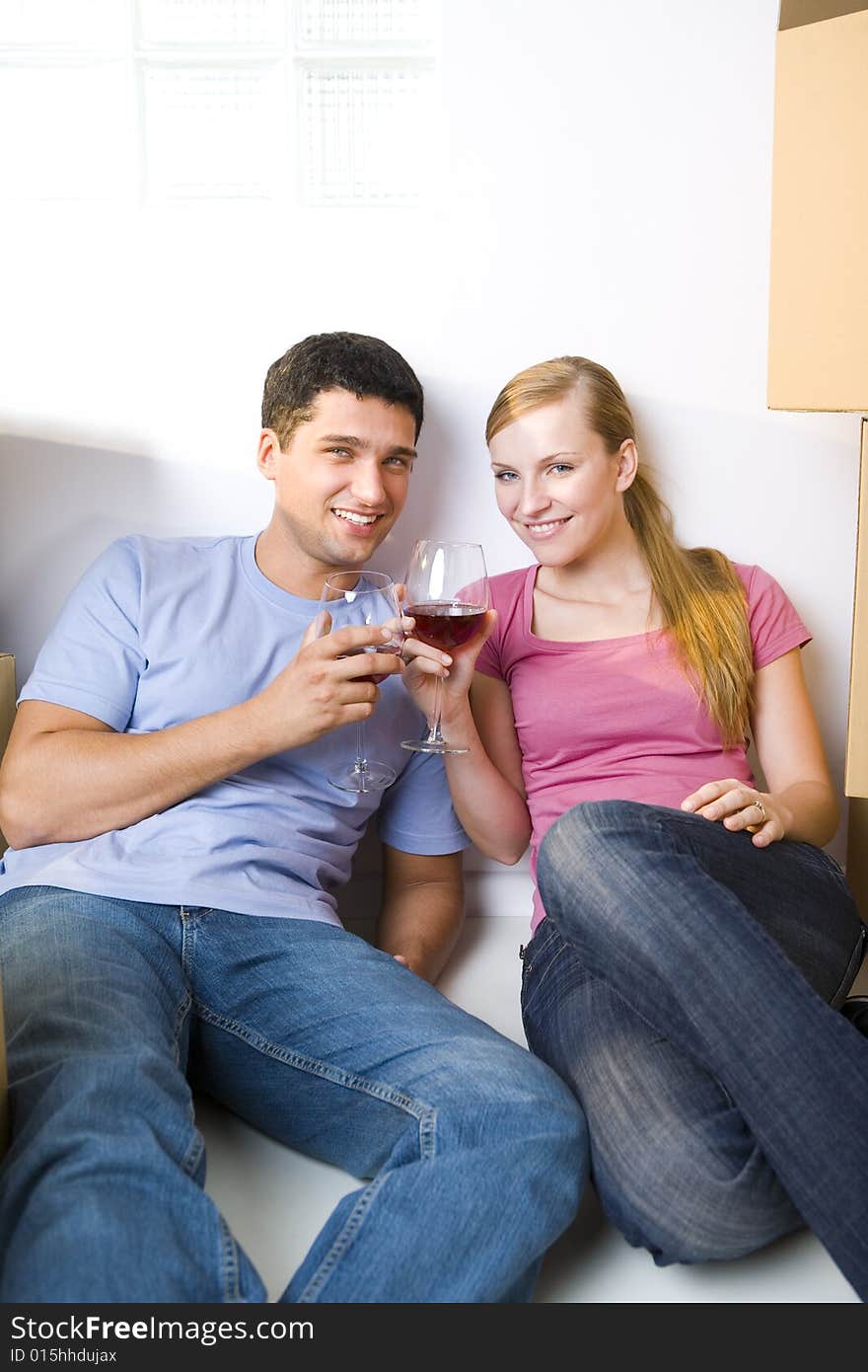 Young couple sitting between cardboard boxes and drinking wine. They're smiling and looking at camera. Front view. Young couple sitting between cardboard boxes and drinking wine. They're smiling and looking at camera. Front view.