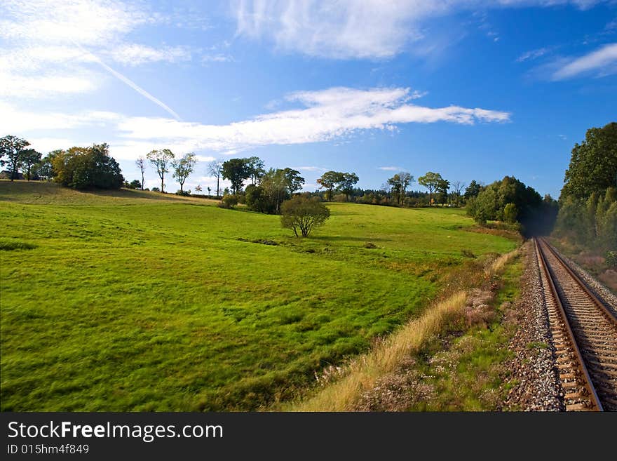 Green meadow with trees next to rail track and blue cloudy sky. Green meadow with trees next to rail track and blue cloudy sky