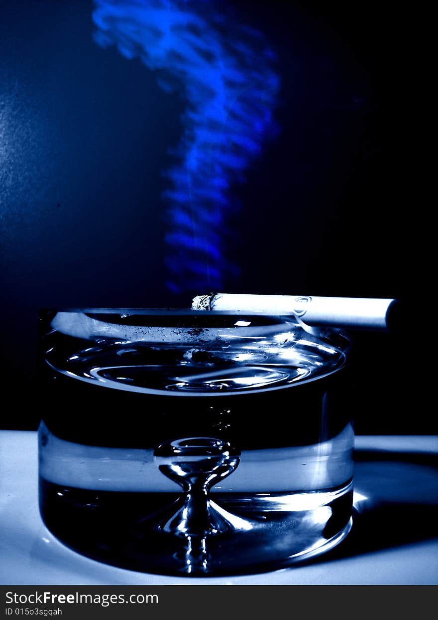 Cigarette with blue smoke on a big glass ash-tray in navy blue color. Cigarette with blue smoke on a big glass ash-tray in navy blue color.