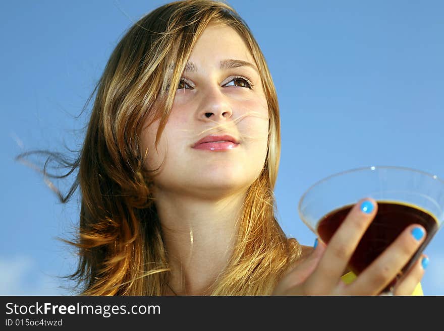 A young beautiful girl with glass of red drink looks far on a background blue sky