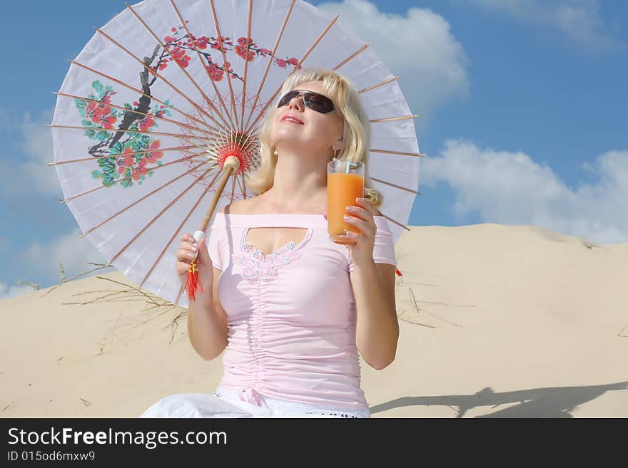 A young blonde woman sits on sand with glass of juice and umbrella. A young blonde woman sits on sand with glass of juice and umbrella