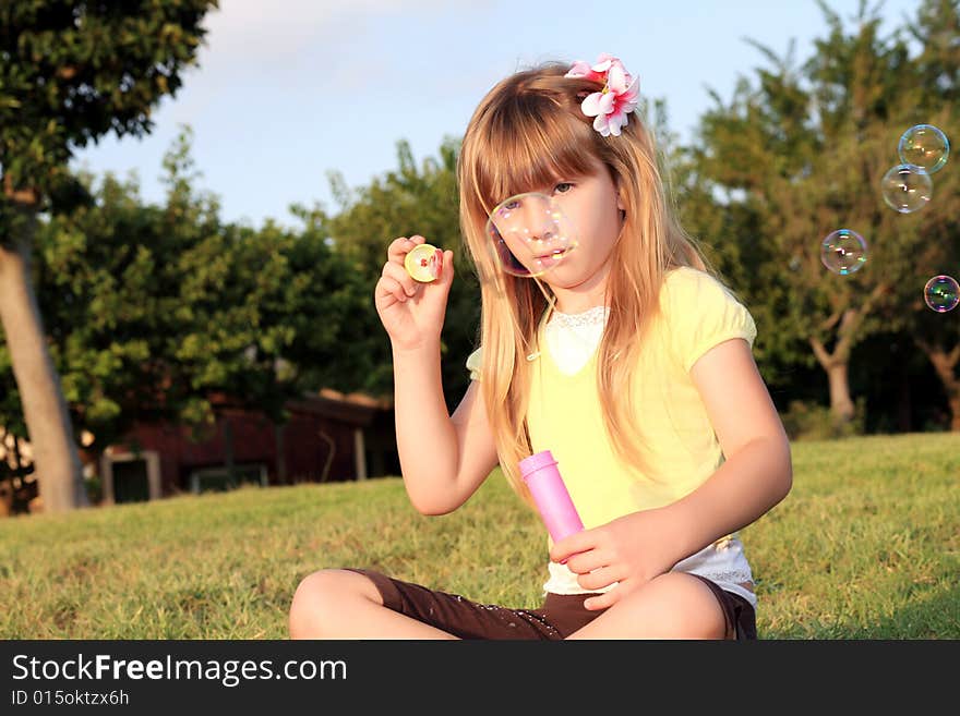 A little beautiful blonde sits on a green grass on a background blue sky and allows soap-bubbles. A little beautiful blonde sits on a green grass on a background blue sky and allows soap-bubbles