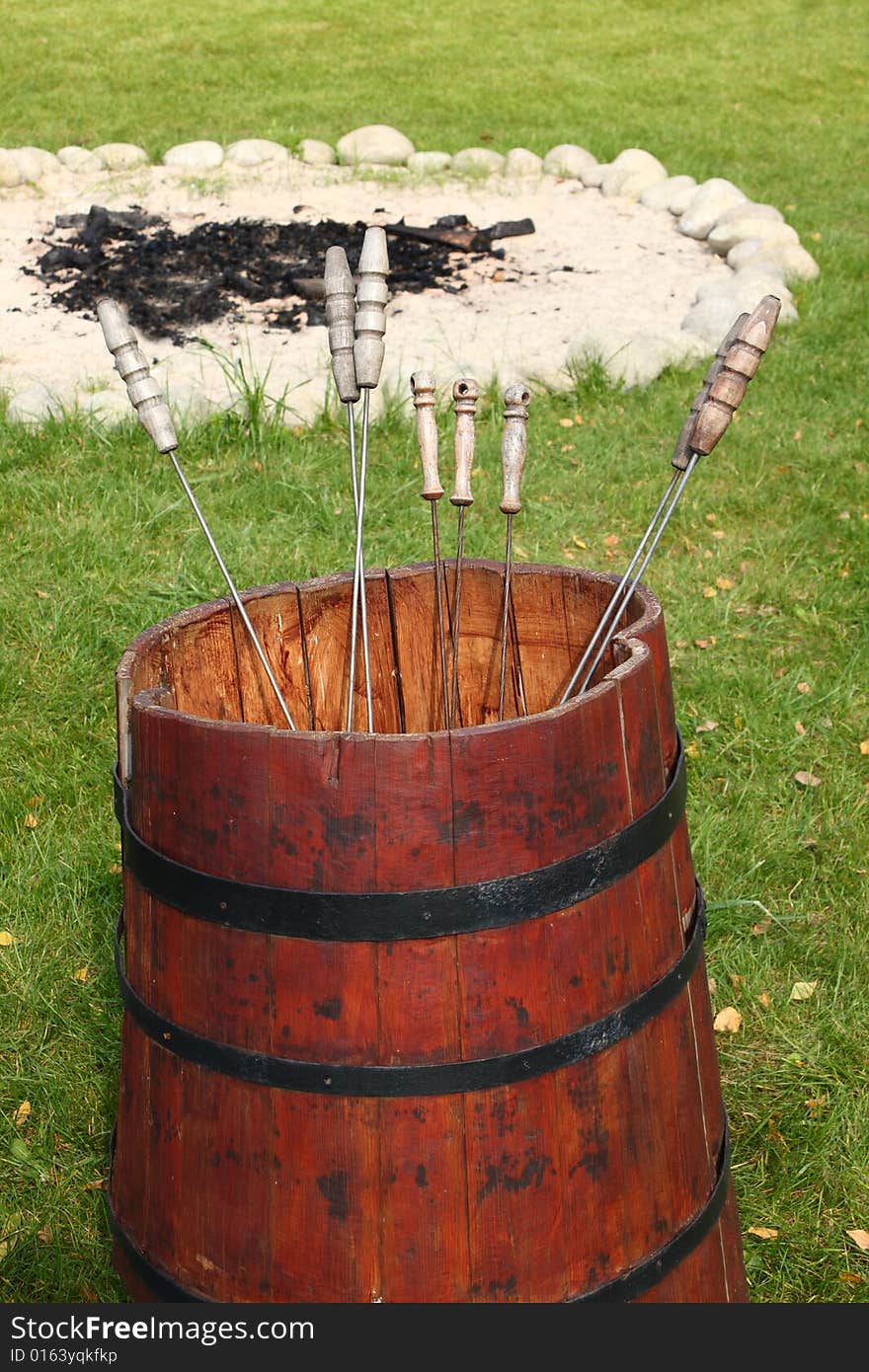 Red barrel with metal skewers. Extinguished campfire in a background.