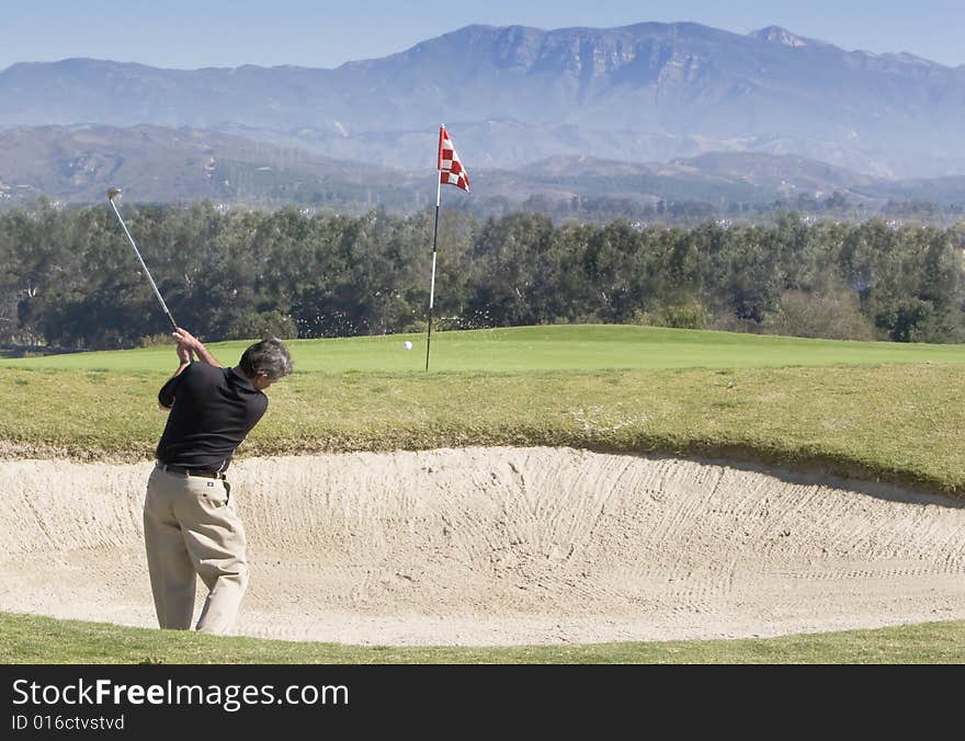 Golfer hitting out of bunker, ball in flight to pin, blue sky and mountains in background. Golfer hitting out of bunker, ball in flight to pin, blue sky and mountains in background