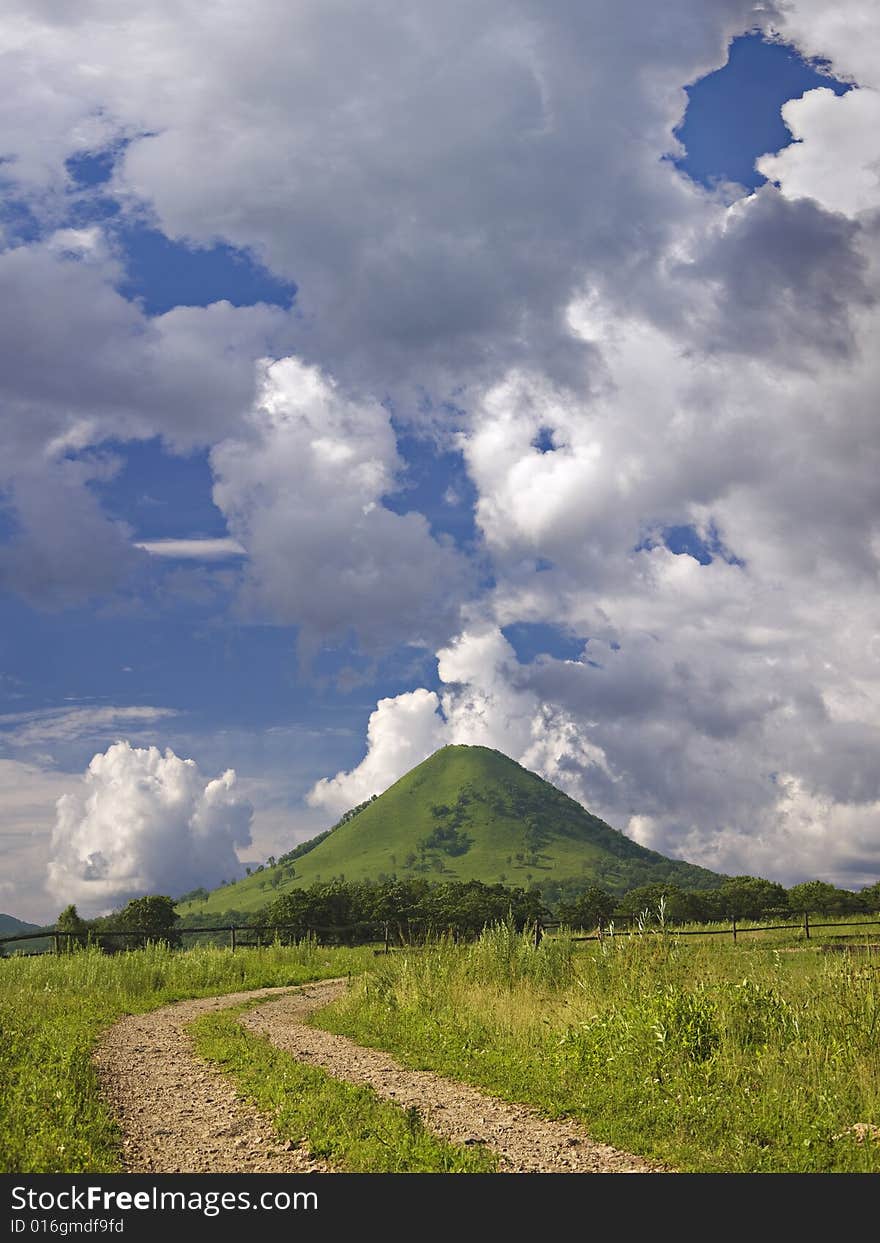 Country road to lonely green mountain under blue sky with quaint clouds in sunny day. Country road to lonely green mountain under blue sky with quaint clouds in sunny day