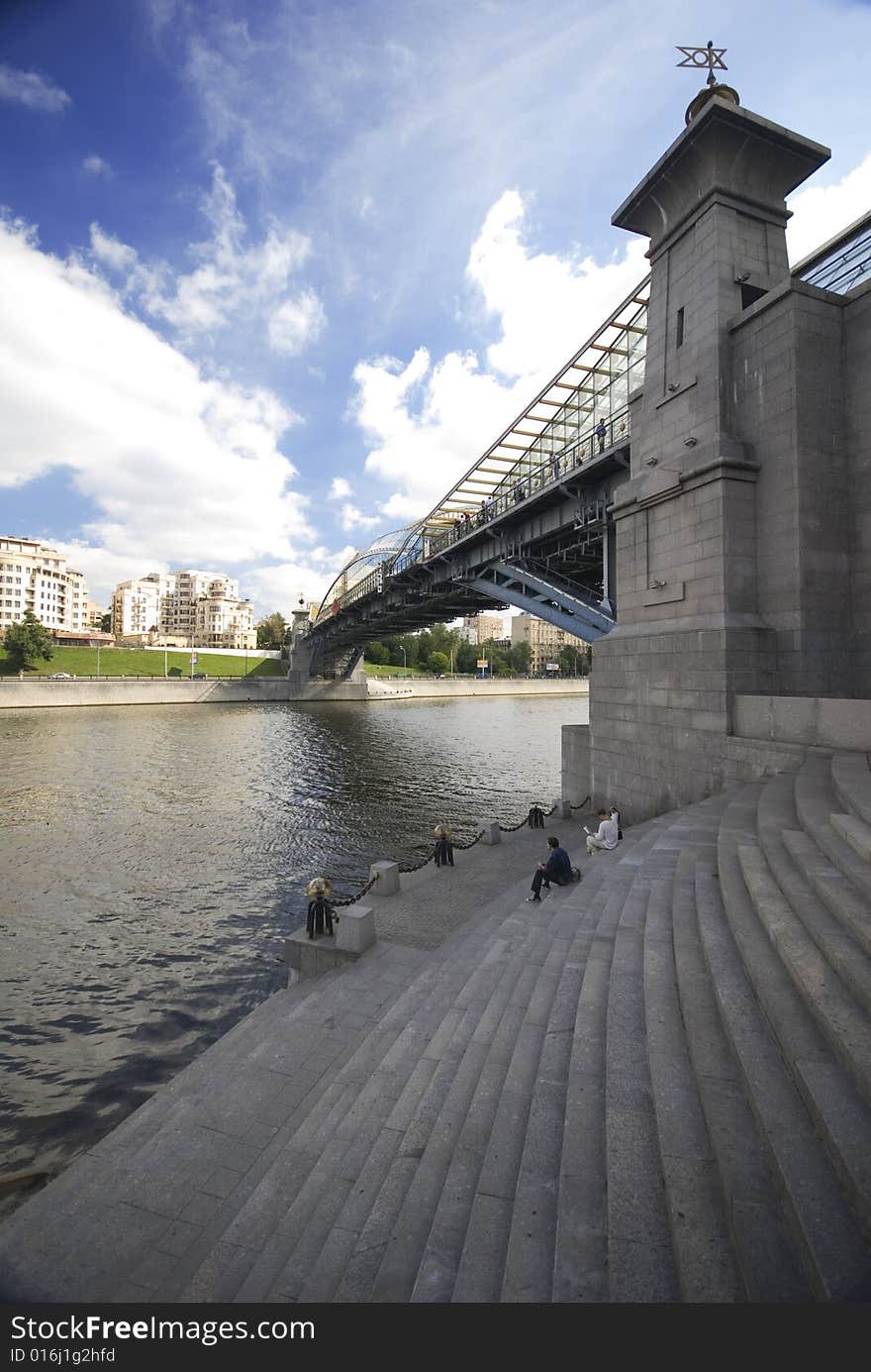 Grey bridge over the river and some people sitting on the bank of this river and clouds in the sky