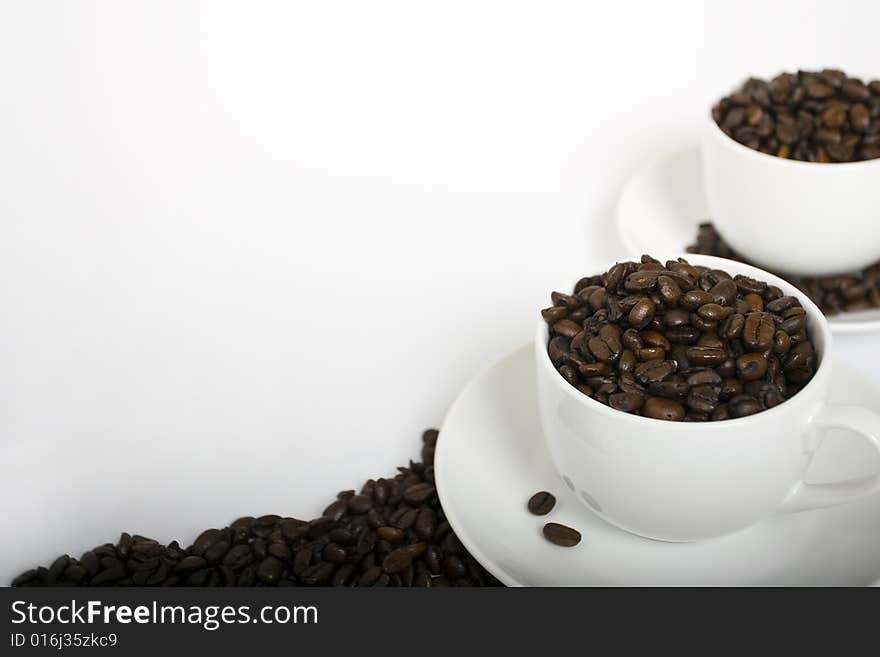 Two coffee cups filled with espresso coffee beans on the right-hand side of a white frame. Beans placed around saucers and a border of beans at the bottom. Two coffee cups filled with espresso coffee beans on the right-hand side of a white frame. Beans placed around saucers and a border of beans at the bottom.