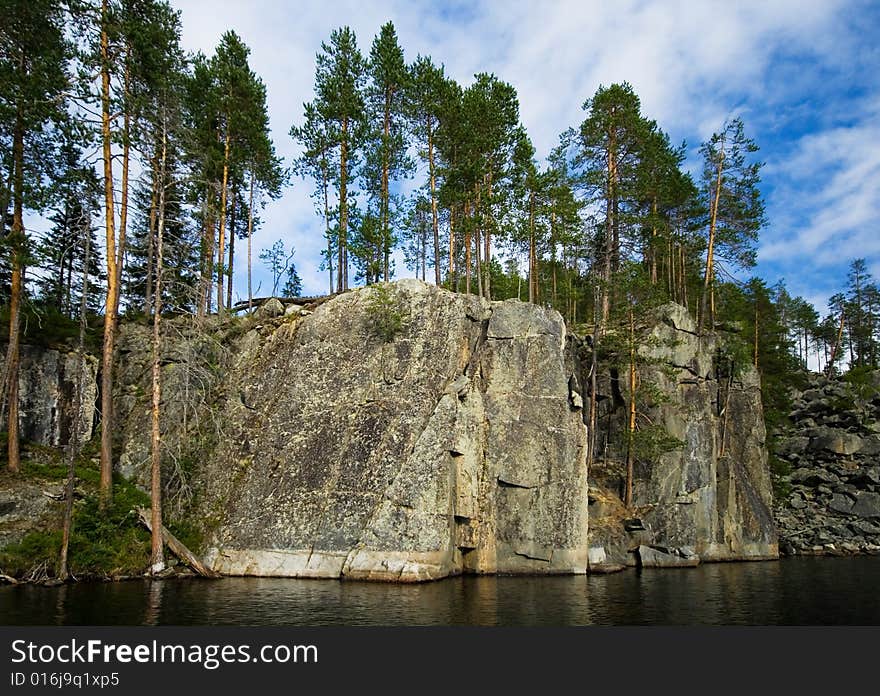 A wild sheer cliff rises from the black water of Karelia Pizanets lake with pine tress on it. A wild sheer cliff rises from the black water of Karelia Pizanets lake with pine tress on it