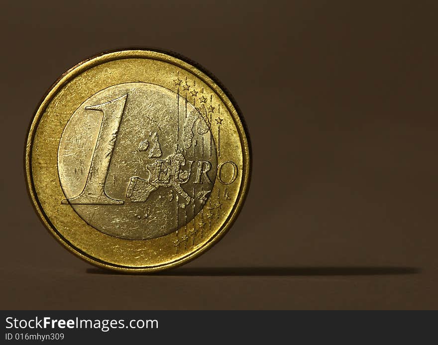 One euro coin balancing on its edge, casting a narrow shadow on the brown background.