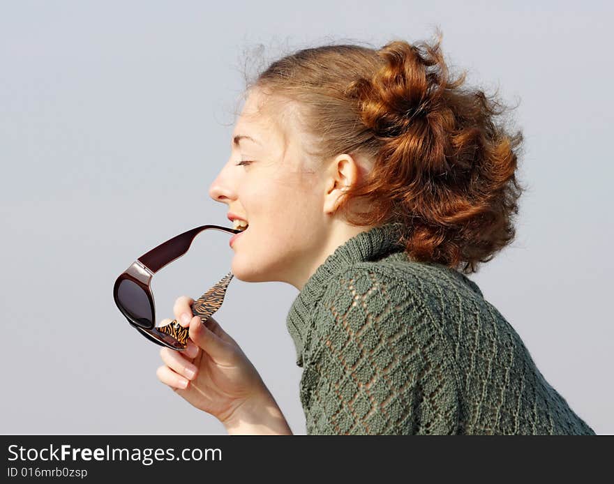 Old-fashioned Young curly girl in a vintage gray knitted dress bites her sunglasses. She smiles slightly and flirts. Blue background.Girl with close-up eyes enjoy the sun. Old-fashioned Young curly girl in a vintage gray knitted dress bites her sunglasses. She smiles slightly and flirts. Blue background.Girl with close-up eyes enjoy the sun.