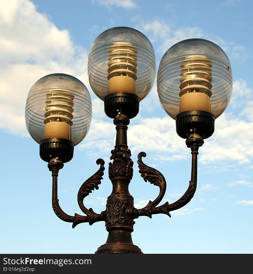 Three ornate street lamps on a lamp post against a blue sky in Romania