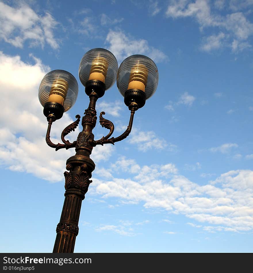 Three ornate street lamps on a lamp post against a blue sky in Romania