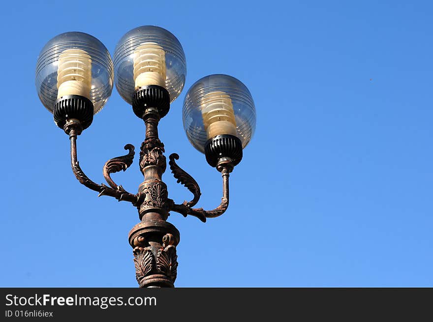 Three ornate street lamps on a lamp post against a blue sky in Romania