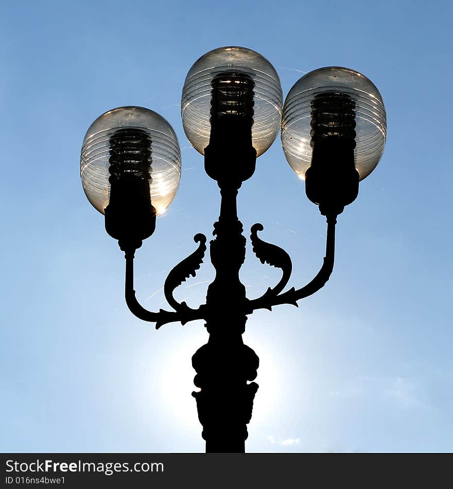 Three ornate street lamps on a lamp post against a blue sky in Romania