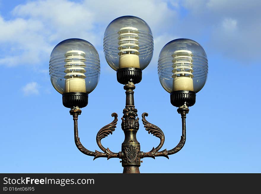 Three ornate street lamps on a lamp post against a blue sky in Romania