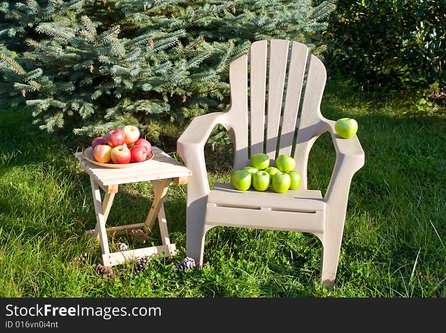 Lounge chair with Granny Smith apples beside an evergreen tree. Wooden table with plate of Gala apples. Lounge chair with Granny Smith apples beside an evergreen tree. Wooden table with plate of Gala apples.