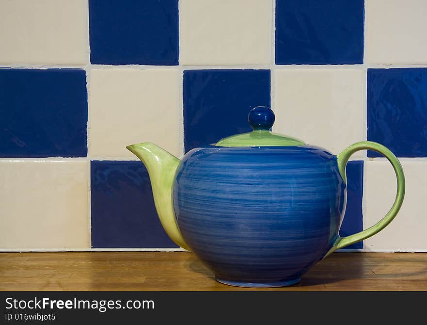 A cheery blue and green teapot stands on a wood counter in front of blue and white tiles. A cheery blue and green teapot stands on a wood counter in front of blue and white tiles.