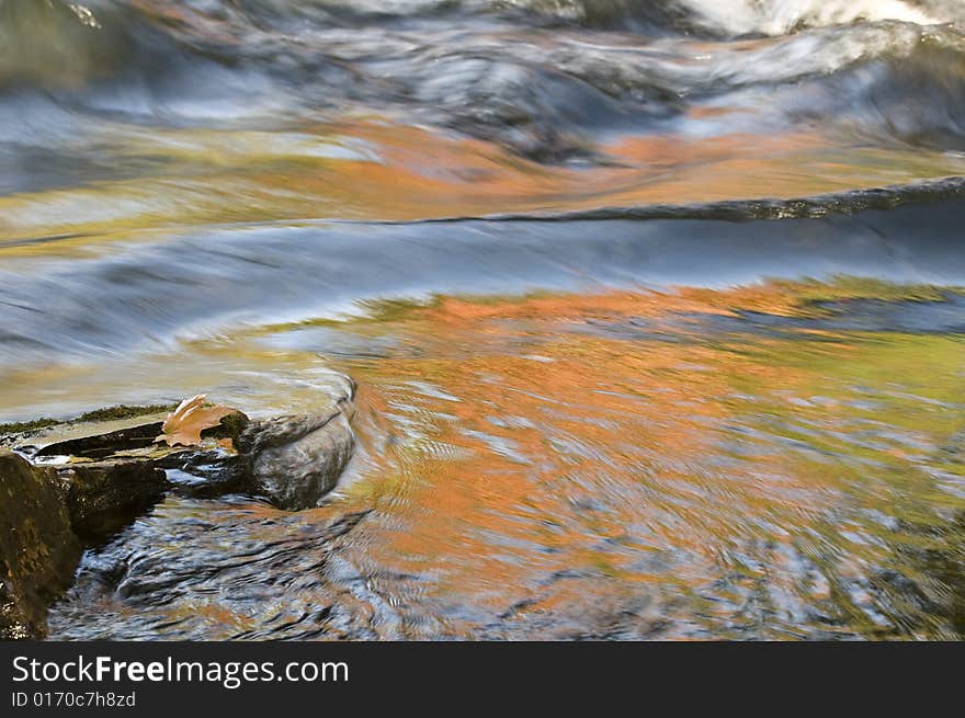 Rapidly moving water is showing the blues and gold colors reflecting with a leaf and rock in the foreground. Rapidly moving water is showing the blues and gold colors reflecting with a leaf and rock in the foreground.