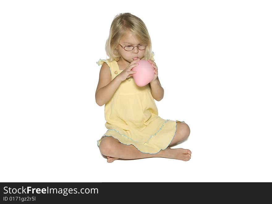 A young girl in a yellow dress sitting down and blowing up a pink balloon. Isolated on a white background. A young girl in a yellow dress sitting down and blowing up a pink balloon. Isolated on a white background