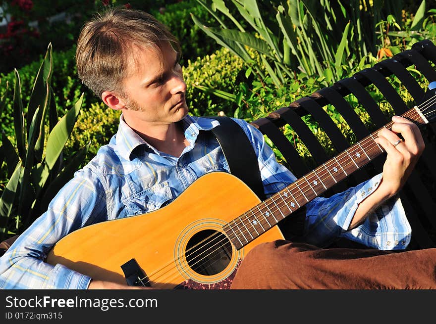 Young man plays guitar outside on a park bench. Young man plays guitar outside on a park bench