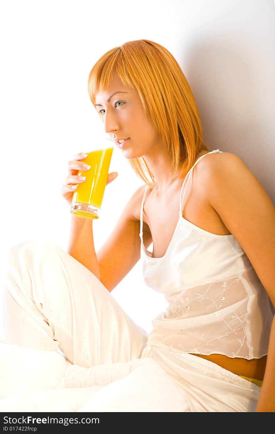 Young red-haired woman dressed pyjamas sitting on bed with glass of juice. She's leaning against a wall. Young red-haired woman dressed pyjamas sitting on bed with glass of juice. She's leaning against a wall.