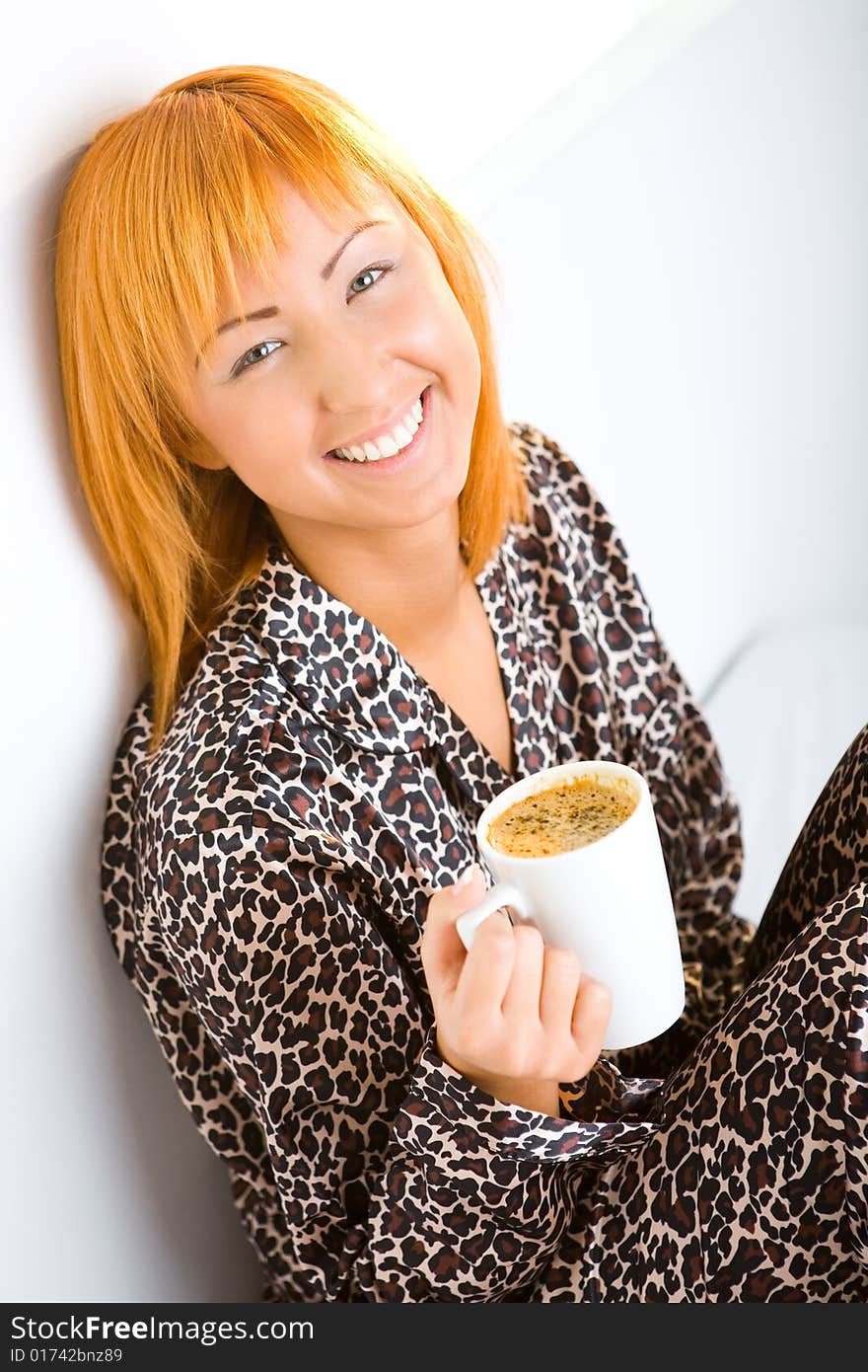 Young red-haired woman dressed pyjamas sitting on bed with cup of coffee. She's leaning against a wall. She's smiling and looking at camera. Young red-haired woman dressed pyjamas sitting on bed with cup of coffee. She's leaning against a wall. She's smiling and looking at camera.