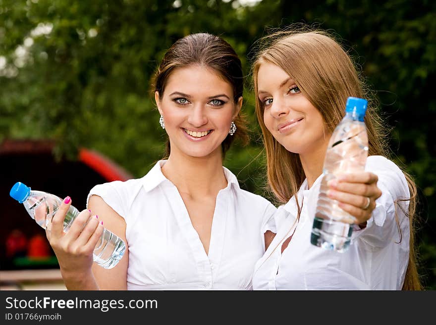 Two inseparable cheerful girlfriends holding bottle of water in summer park. Two inseparable cheerful girlfriends holding bottle of water in summer park