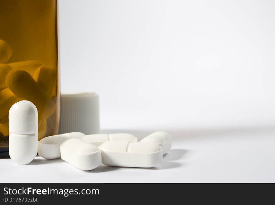 A group of pills on the lower left-hand side of a white frame with one standing up next to a brown bottle. A group of pills on the lower left-hand side of a white frame with one standing up next to a brown bottle