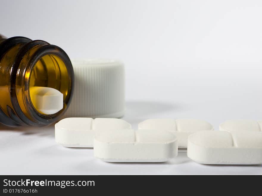 The mouth of a bottle on the left-hand side of a white background with a row of pills arranged along the bottom of the frame. The mouth of a bottle on the left-hand side of a white background with a row of pills arranged along the bottom of the frame