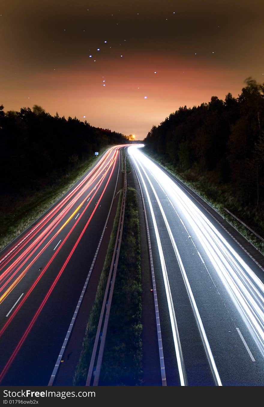 Night traffic leaving read and white trails along a highway leading to a constallation of stars in a moody sky,. Night traffic leaving read and white trails along a highway leading to a constallation of stars in a moody sky,