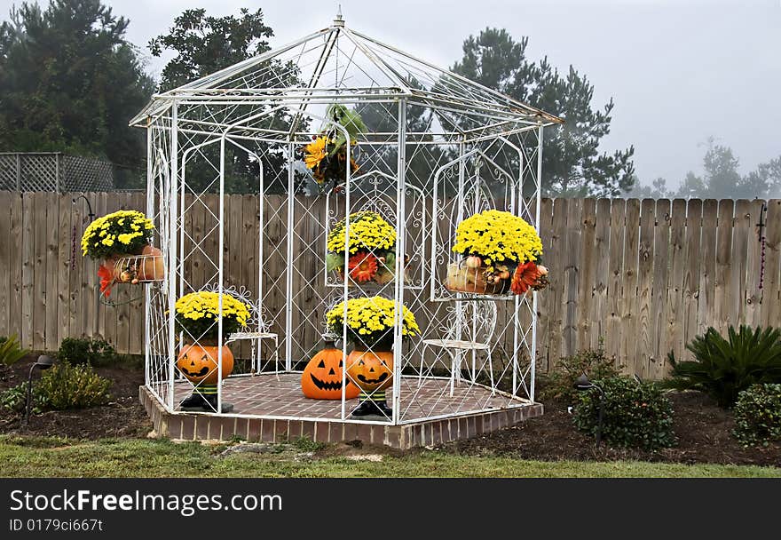 A white iron gazebo decorated for fall with mums, pumpkins and jack-o-laterns overcast by a hazy sky. A white iron gazebo decorated for fall with mums, pumpkins and jack-o-laterns overcast by a hazy sky.