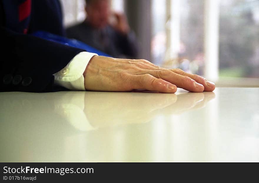 Close up of hand of business man resting on a table, with person out of focus talking on phone in background. Close up of hand of business man resting on a table, with person out of focus talking on phone in background