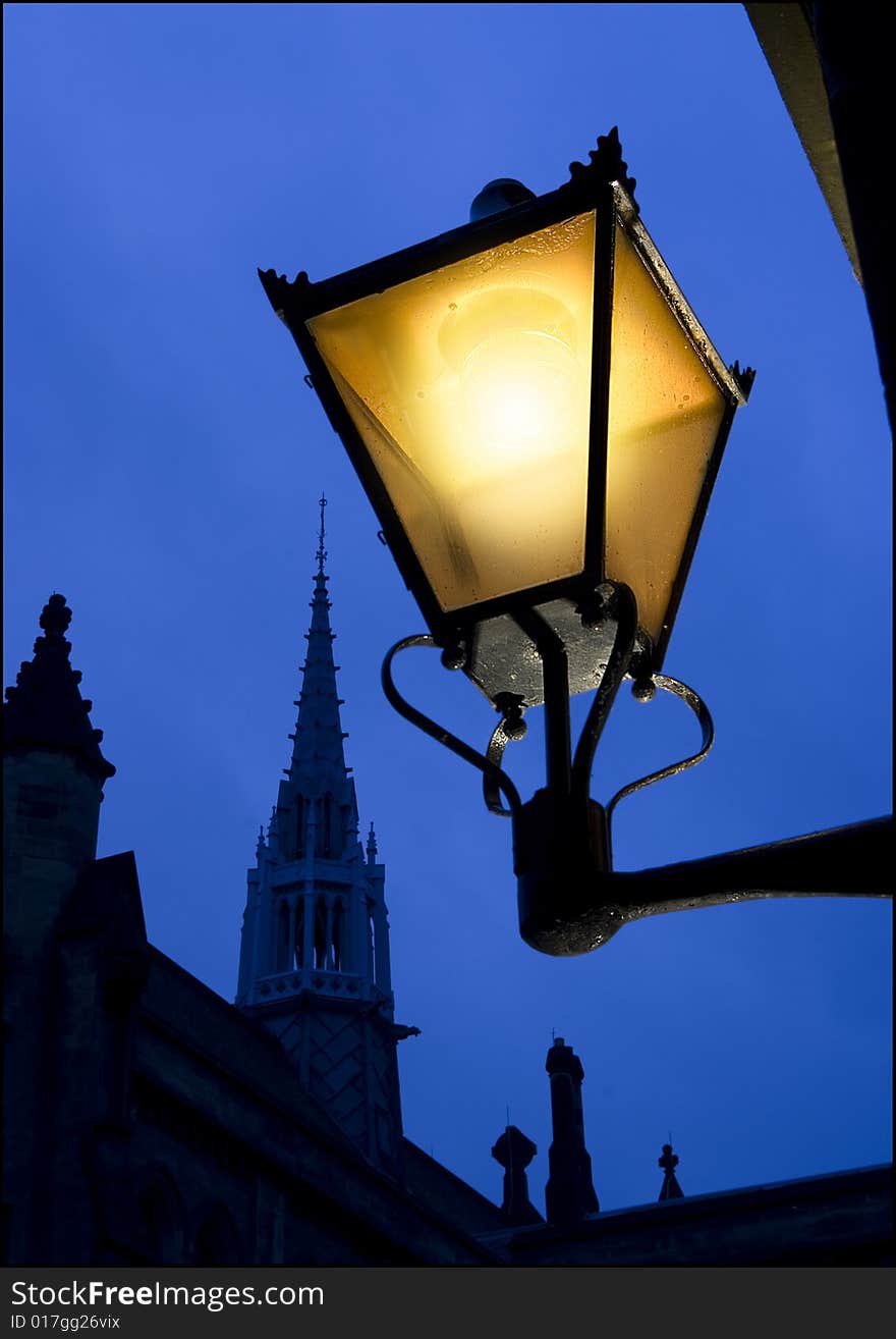 Traditional street lamp with glowing yellow light contrasted with blue sky and dark buildings. Concept for warmth of light and feeling of home and security. Traditional street lamp with glowing yellow light contrasted with blue sky and dark buildings. Concept for warmth of light and feeling of home and security.